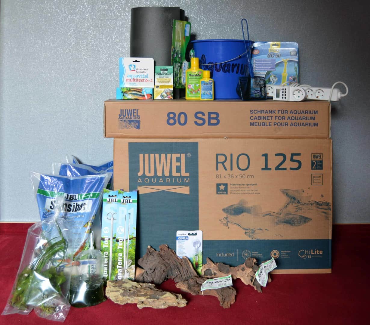 Einkaufsliste Aquarium und Unterschrank im Karton, davor Eimer, Pflegemittel, Bodengrund, Pflanzen und Wurzeln von der Einkaufsliste.